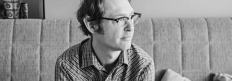 A man with glasses and a plaid shirt sits on a sofa, looking pensively into the distance. The room is softly lit, creating a calm and introspective atmosphere. Black and white.