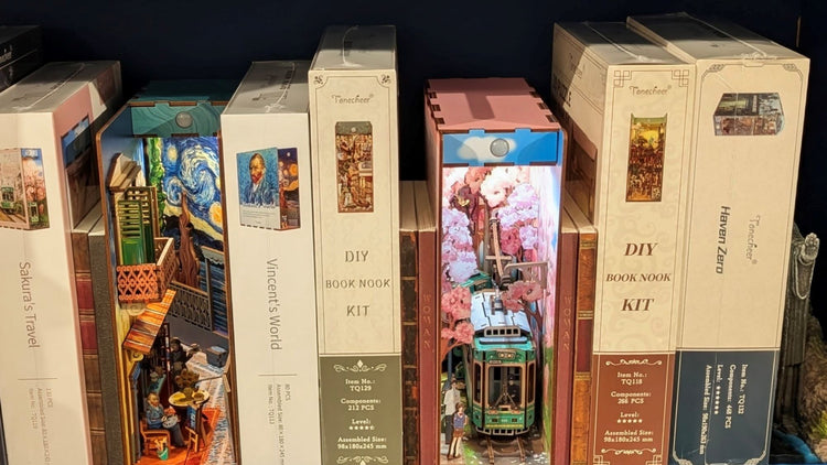 Several DIY book nook kits are displayed on a shelf, with some completed examples showing miniature scenes, including cherry blossoms with a tram, a marketplace, and Van Gogh’s Starry Night.
