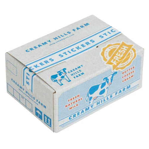 A rectangular box labeled "Japanese Mini Sticker Box Creamy Hills" from Blue Dog Posters, featuring blue and red illustrations of cows and a gold "Fresh" seal; the box contains sticker sheets.
