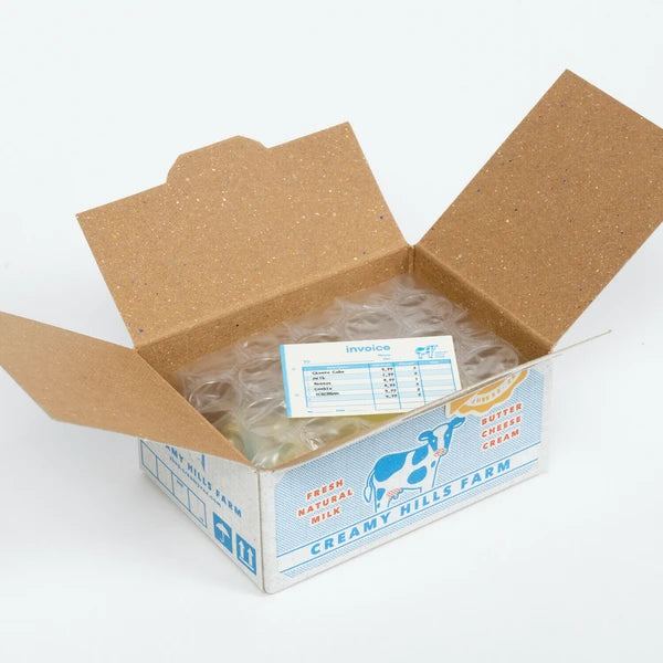 An open cardboard box labeled "Japanese Mini Sticker Box Creamy Hills" by Blue Dog Posters with a cow illustration contains an invoice and bubble wrap inside. The flaps are open, revealing the contents.