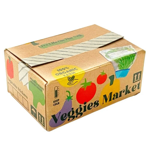 A small brown box labeled "Japanese Mini Sticker Box Veggies Market" by Blue Dog Posters features colorful veggie illustrations, a "100% Organic" sticker, and a clear window displaying green leafy stickers inside.