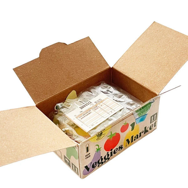 An open cardboard box labeled "Veggies Market" with colorful veggie art reveals a sealed Japanese Mini Sticker Box by Blue Dog Posters, safely packed in bubble wrap.