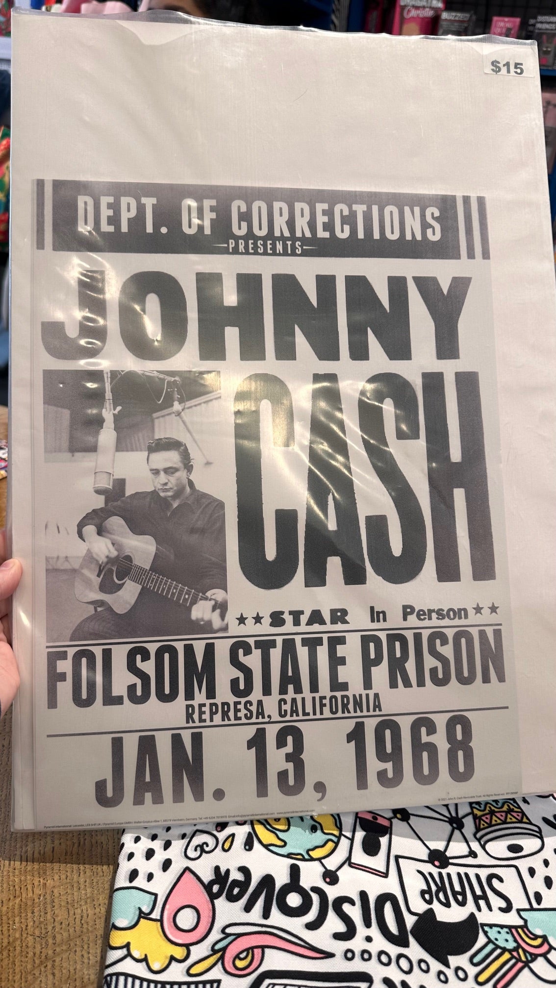 A person is holding a vintage "Johnny Cash Folsom State Prison" poster by Blue Dog Posters, featuring the text "Dept. of Corrections presents Johnny Cash - Star in Person - Folsom State Prison, Represa, California - Jan. 13, 1968." The poster depicts a black and white image of Johnny Cash playing a guitar. Price tag reads $15.