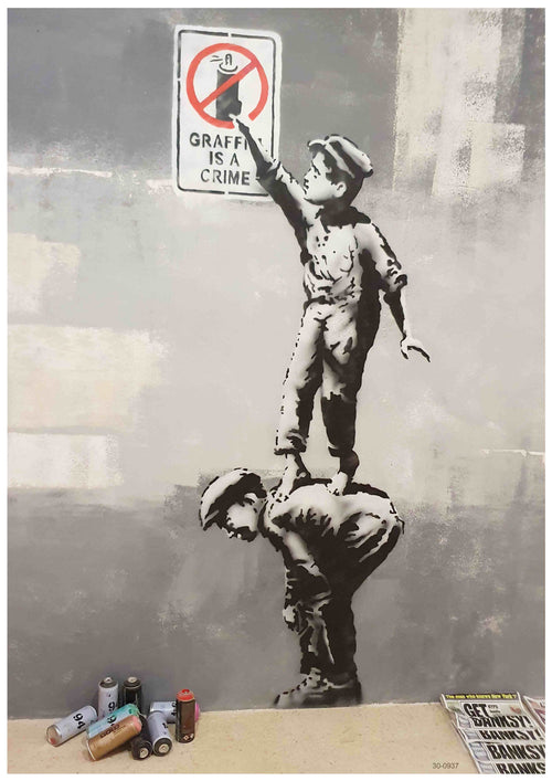 A mural from the 'Banksy Graffiti Is a Crime' collection by brand Y depicts two boys in black and white. One boy is crouched, serving as a step for the other, who stands on his back, reaching to paint over a "Graffiti is a crime" sign. Various spray paint cans and stencils lie on the ground next to them, creating an iconic Banksy Poster effect perfect for A3 Satin Paper prints.