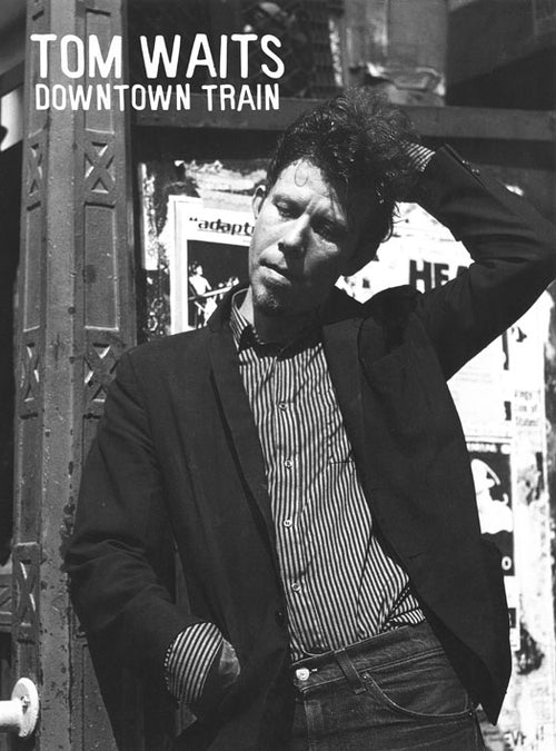A black and white image of a man in a striped shirt and blazer, standing against a rough urban backdrop adorned with posters. The man is slightly hunched, touching his head with a pensive expression. "TOM WAITS" and "DOWNTOWN TRAIN" are prominently displayed at the top. Perfect for any music collection, this product is listed as Tom Waits - Downtown Train by MUS 212.