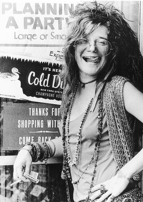 A woman with long hair and glasses smiles while leaning against a wall. She is adorned with a beaded necklace, bracelets, a crocheted vest, and holding a cigarette. Behind her is the "Planning a Party" sign advertising Cold Duck wine, all printed on Premium 150gsm gloss paper for the Janis Joplin Planning a Party poster by MUS 096.
