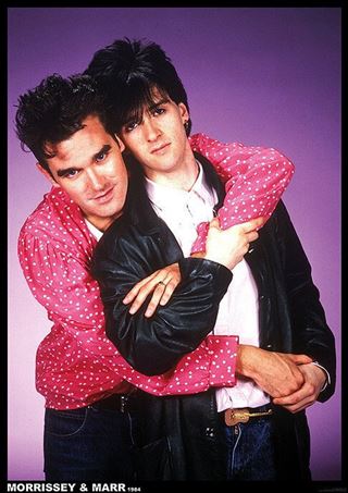 Two men with dark hair and serious expressions pose against a purple backdrop. One, in pink polka dots, hugs the other, who wears a black leather jacket. Featured: The Smiths Morrissey & Marr 1984 Colour by MUS 279.
