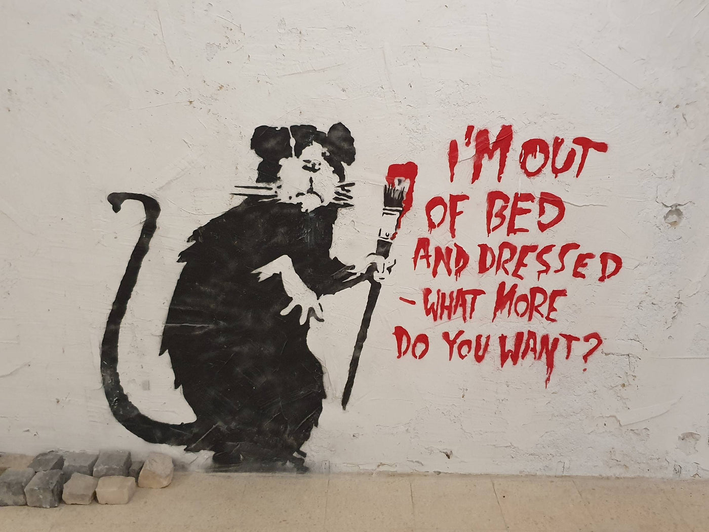 The Banksy Out of Bed Rat poster by Y features a graffiti artwork on a rough white wall, depicting a black rat standing on its hind legs and holding a red paintbrush. Red text next to the rat reads, "I'M OUT OF BED AND DRESSED - WHAT MORE DO YOU WANT?" Printed on high-quality 250gsm Satin Paper, this A3 poster captures every detail perfectly.