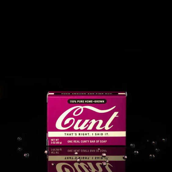 A magenta-packaged bar of soap set against a black background. The vintage-style box boldly displays the word "Cunt" in large cursive font, accompanied by the slogans "That's right. I said it." and "One real cunty bar of soap." This daring creation is part of the Cunt Triple Milled Boxed Bar Soap line from Blue Dog Posters.