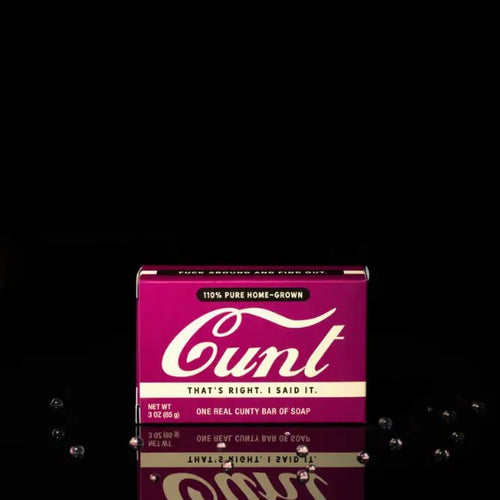 A magenta-packaged bar of soap set against a black background. The vintage-style box boldly displays the word "Cunt" in large cursive font, accompanied by the slogans "That's right. I said it." and "One real cunty bar of soap." This daring creation is part of the Cunt Triple Milled Boxed Bar Soap line from Blue Dog Posters.