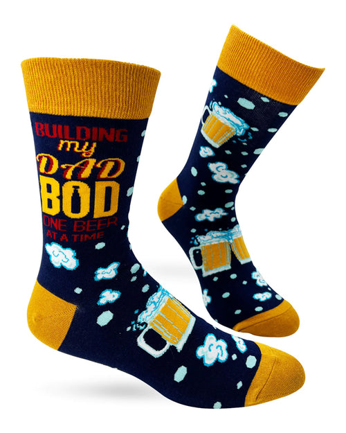 Check out the One Beer at a Time Socks for men by Blue Dog Posters. This pair of navy blue socks features a humorous design with beer mugs and foam, accompanied by the text "Building my Dad Bod one beer at a time" in red, yellow, and white. These comfy socks fit men's shoe sizes 7 to 12 and come with stylish yellow cuffs, heels, and toes.
