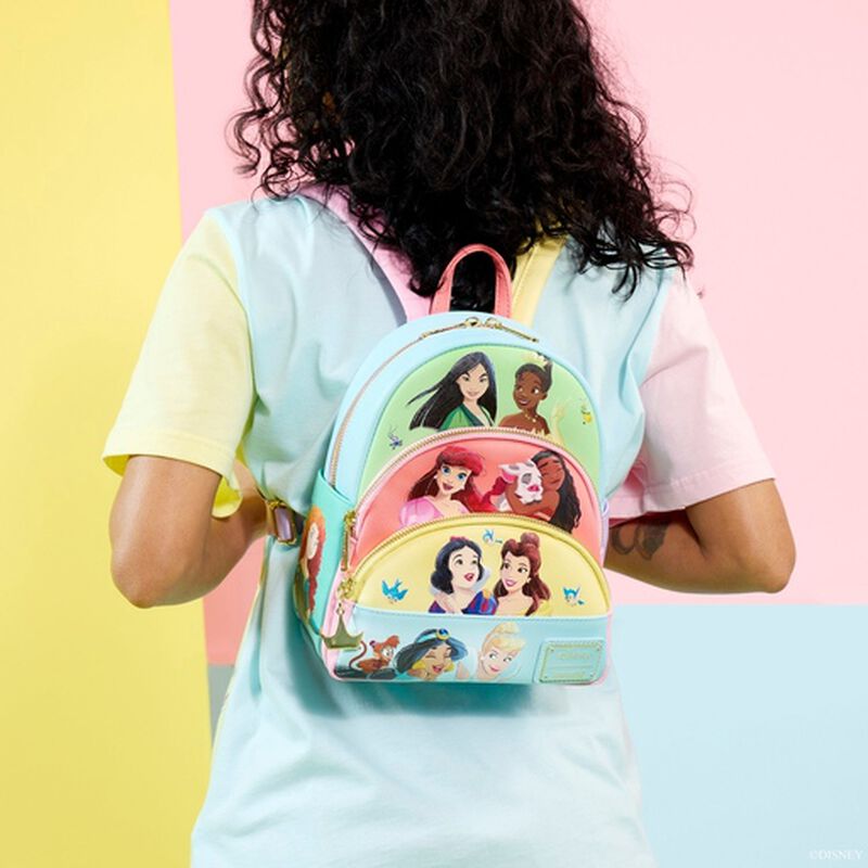 A person with curly hair, wearing a pastel-colored shirt and facing away from the camera, is showcasing a vibrant Disney Princess Collage Mini Backpack by Blue Dog Posters. The background features sections of pastel pink and yellow.