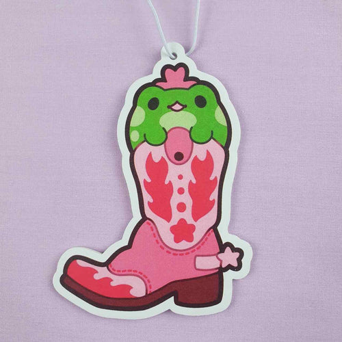 The Cowboy Froggy Watermelon Air Freshener by Blue Dog Posters is a vibrant and decorative tag that showcases a whimsical pink cowboy boot with striking red accents. Nestled inside the boot is an adorable green and pink creature with round eyes and a small beak. The soft lavender background enhances its charm, while it also serves as an air freshener, emitting a delightful watermelon fragrance.