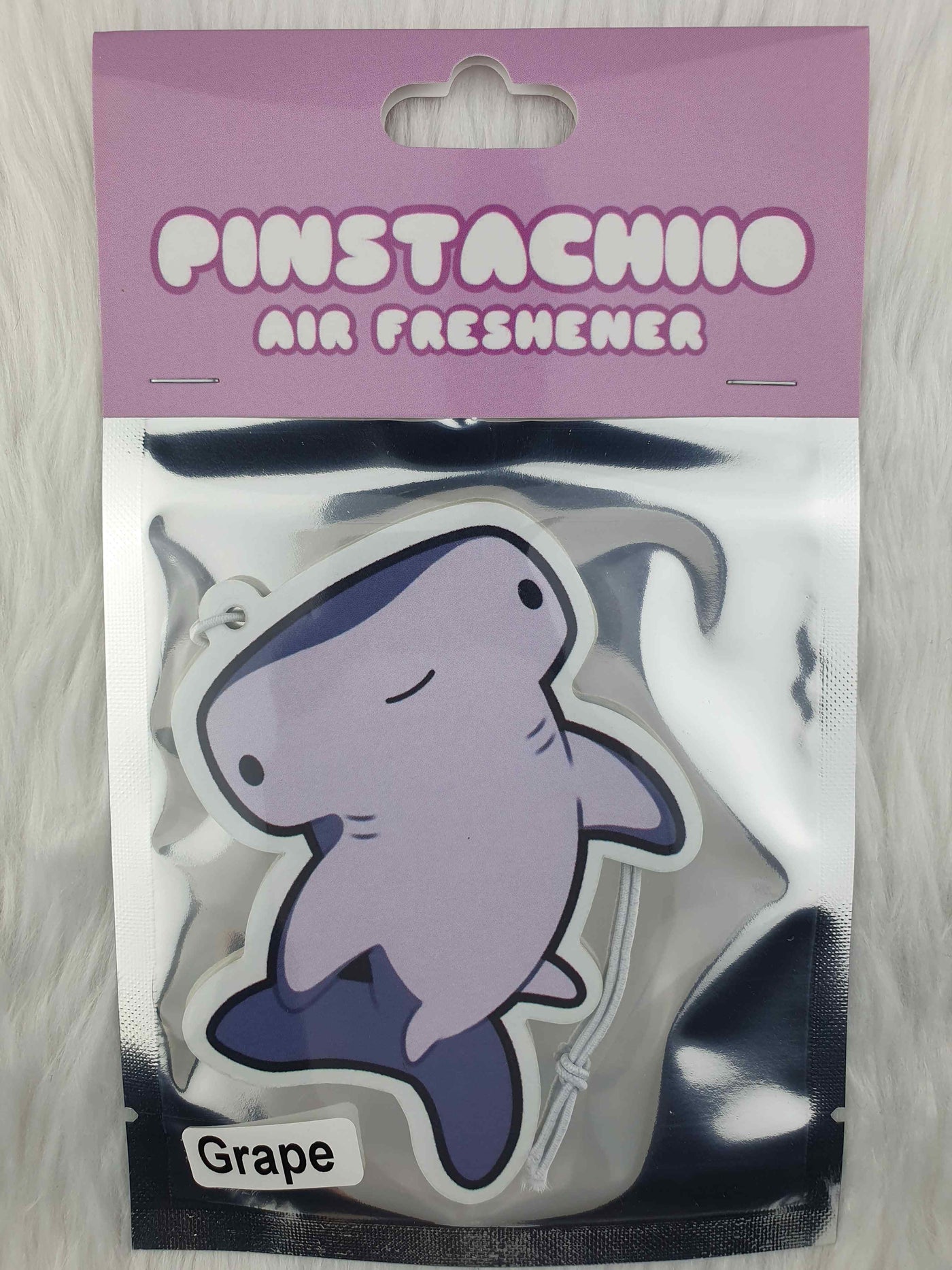 A packaged air freshener featuring a charming cartoon hammerhead shark design is visible. The packaging reads "Pinstachiio Air Freshener" at the top and "Grape" at the bottom, highlighting its grape fragrance. The background showcases soft, fluffy white fabric, while a small "Made in Australia" emblem adds a touch of charm. This product is named Hammerhead Shark Grape Air Freshener by Blue Dog Posters.