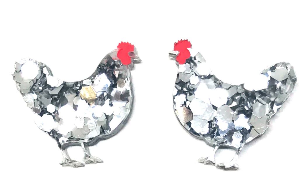 Two handmade Hen Glitter Stud Earrings in silver, shaped like chickens facing each other, each featuring a small red comb on top. These earrings have a sparkly, reflective surface, creating a festive and shiny appearance. From Blue Dog Posters' latest collection, these pieces add a touch of charm to any setting.