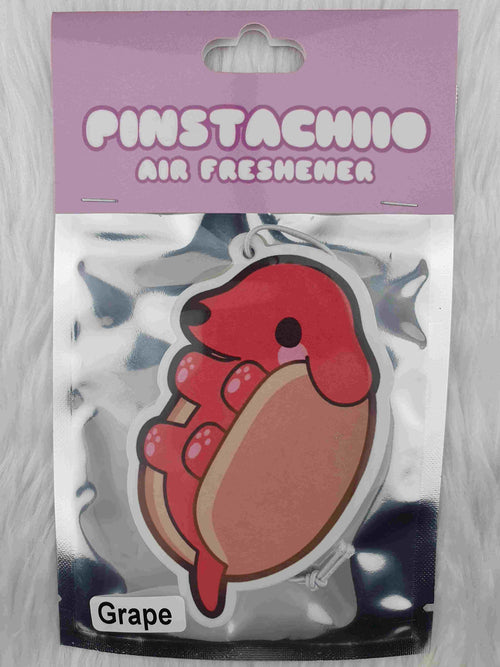 A packaged air freshener featuring a cute cartoon design of a red dachshund nestled in a bun, resembling a hot dog. The top of the package has a lavender header with "Pinstachiio Air Freshener" in playful white text and a label indicating the grape fragrance at the bottom. Made in Australia by Blue Dog Posters.