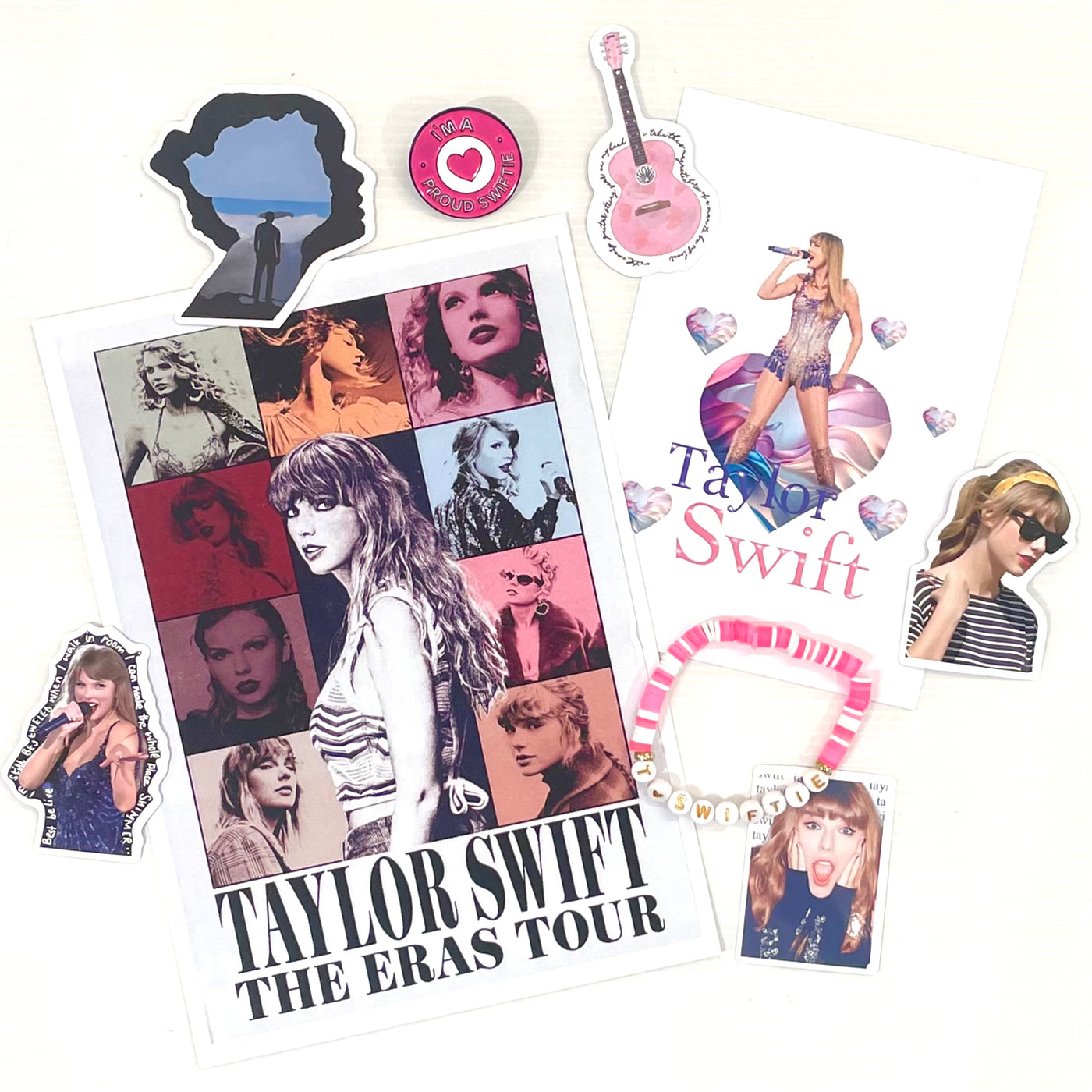 The Blue Dog Posters Mystery Goodie Bag - Taylor Swift includes an Eras Tour poster, Taylor stickers, a pink beaded bracelet, postcard, guitar sticker, and “The Eras Tour” button displayed on a white surface.
