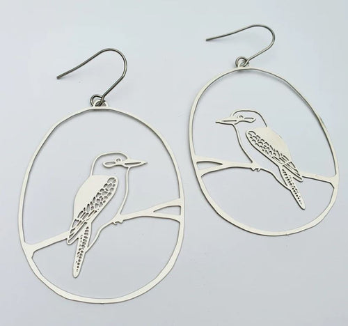 The Denz Kookaburra Dangles Silver Earrings by Blue Dog Posters are a pair of lightweight statement earrings with open oval frames showcasing intricately cut-out designs of Kookaburras perched on branches. Made from recycled stainless steel, the detailed cutouts depict the birds in profile view. The earrings feature simple hooks for easy wearing.