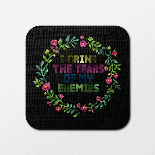 The Tears of My Enemies Coaster by Blue Dog Posters features cross-stitched art on black fabric with colorful flowers forming a circle, surrounding the phrase "I DRINK THE TEARS OF MY ENEMIES" in green, blue, pink, and yellow letters.