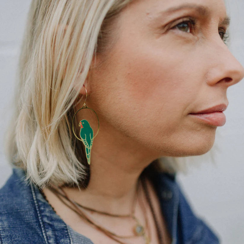 A close-up side profile of a person with blonde hair wearing a large Parrot in Green Denz earring by Blue Dog Posters. The person is dressed in a denim jacket and layered necklaces, looking off into the distance and showcasing their unique Australiana accessory.