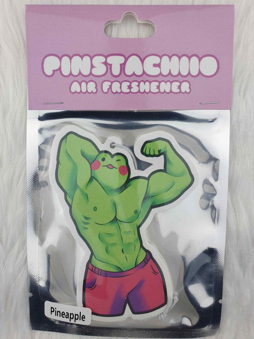 A packaged air freshener featuring a muscular green frog in red shorts flexing its muscles. The packaging, labeled "Muscle Froggy Pineapple Air Freshener - Pinstachiio," stands out on a lavender background with "Pineapple" in bold black text on a white label. Enjoy the pineapple fragrance, proudly made in Australia by Blue Dog Posters.