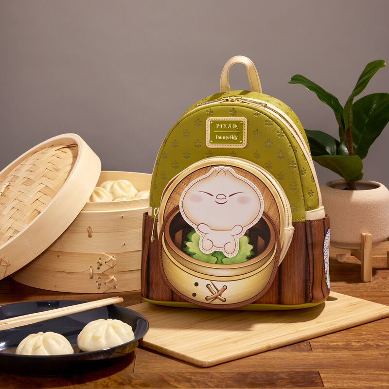 A light green Bao Bamboo Steamer Mini Backpack by Blue Dog Posters, made from vegan leather and featuring an illustration of a dumpling character sitting in a bamboo steamer, is displayed on a wooden table. Behind it are bamboo steamers with buns, a small potted plant, and a plate with chopsticks and steamed buns.