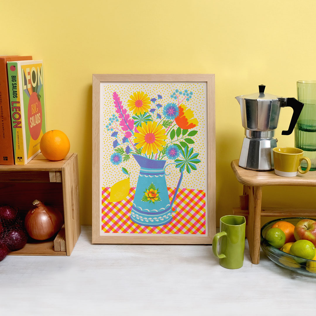 A vibrant still-life scene featuring "Summer Bloom Riso" by Printer Johnson from Blue Dog Posters, with a colorful floral illustration in a frame, accompanied by a moka pot, fruits, and books on wooden shelves. The Risograph print enhances the cheerful yellow background and beautifully complements the vivid colors of the flowers and kitchen items.