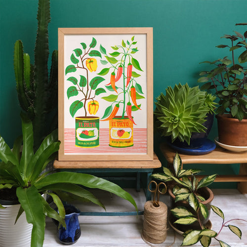 The Chilli Peppers Riso Print by Printer Johnson from Blue Dog Posters showcases a lively A3 Risograph design with colorful El Pato sauce cans nestled among chili plants, succulents, and a cactus on a platform against a teal wall. Crafted with eco-friendly soy-based inks, the print also features nearby scissors and twine for an added creative flair.