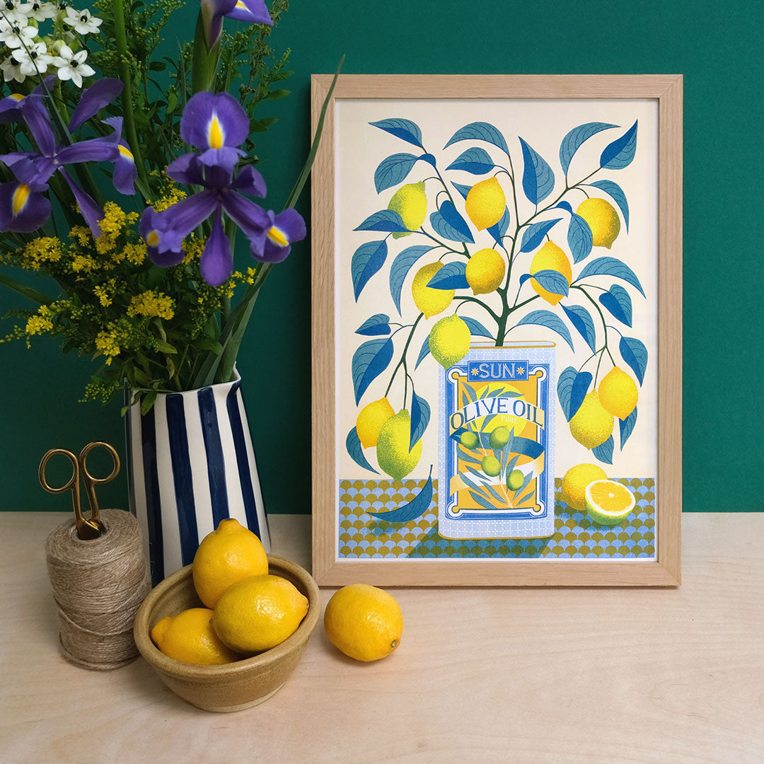 A framed illustration titled "Lemon Tree Riso Print" by Printer Johnson, from Blue Dog Posters, sits on a shelf. It features a lemon tree in a blue "Sun Olive Oil" can and is crafted as a vibrant Risograph print. Nearby, a bowl of lemons and twine with scissors add to the scene. Against the green wall, purple and yellow flowers are displayed in a striped vase, highlighting the eco-friendly soy-based inks used in the artwork.