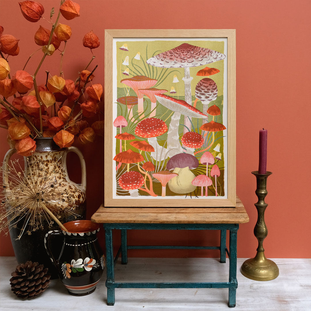 A framed *Fungi Riso Print by Printer Johnson* from Blue Dog Posters, featuring vibrant depictions of various mushrooms, sits on a wooden stand. Surrounding it are vases with orange flowers, a pine cone, and a red candle in a brass holder. The warm coral color serves as the background.