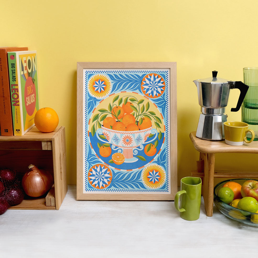 A colorful kitchen scene features the "Orange Bowl Riso Print" by Printer Johnson for Blue Dog Posters, showcasing a framed artwork of oranges in a bowl with geometric patterns. A coffee maker, mugs, and fresh fruits are arranged on wooden shelves nearby. The environment is enriched with cookbooks and vibrant produce against a warm yellow backdrop, enhanced by eco-friendly soy-based inks.