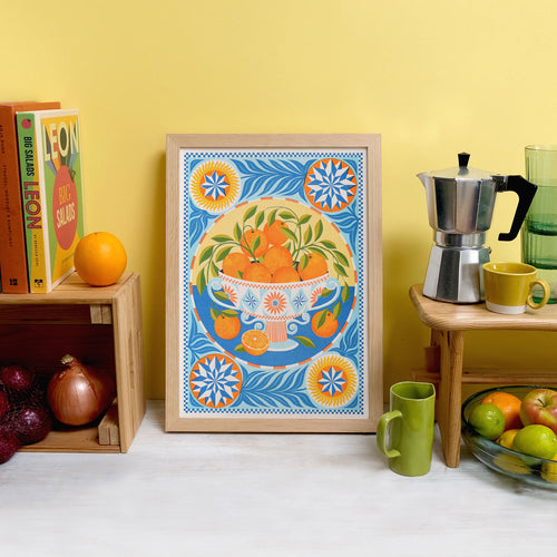 A colorful kitchen scene features the "Orange Bowl Riso Print" by Printer Johnson for Blue Dog Posters, showcasing a framed artwork of oranges in a bowl with geometric patterns. A coffee maker, mugs, and fresh fruits are arranged on wooden shelves nearby. The environment is enriched with cookbooks and vibrant produce against a warm yellow backdrop, enhanced by eco-friendly soy-based inks.