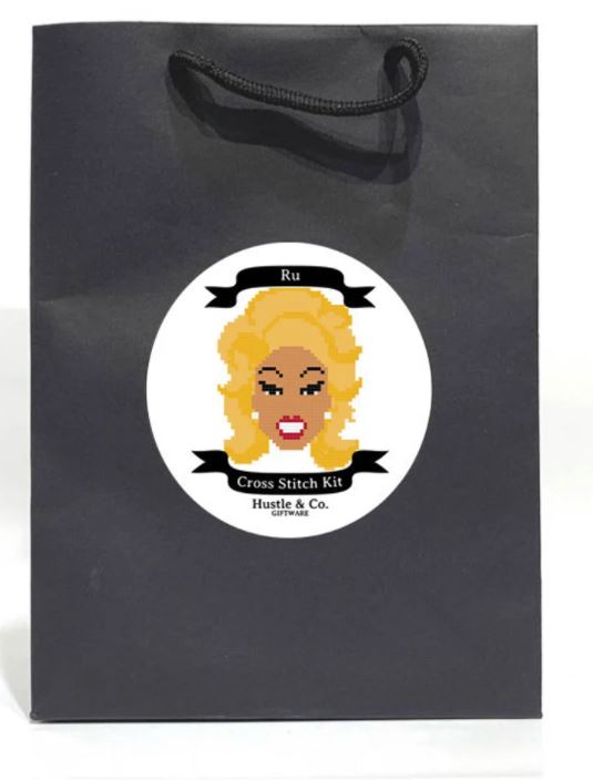 A black bag with a circular label showcasing a pixel art image of Ru Paul, featuring blonde hair. The label reads "Hustle and Co. Cross Stitch - Ru Paul" and "Blue Dog Posters," part of the Cool Gifts Collection. It includes an embroidery hoop, and the bag has a black cord handle at the top.
