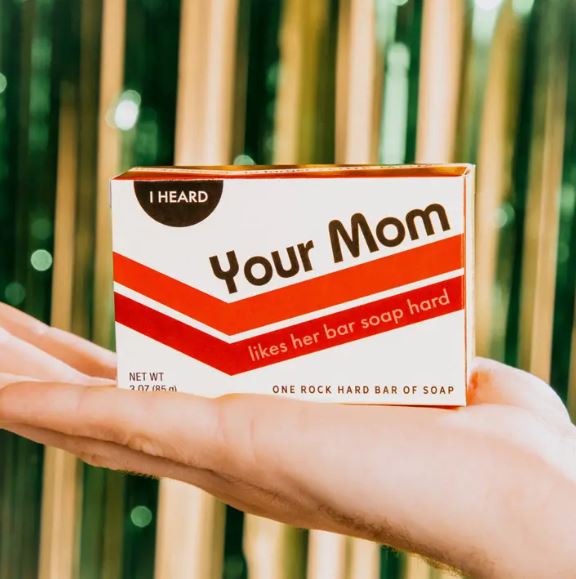 Hands hold a bar of Blue Dog Posters' Your Mom Boxed Bar Soap, packaged in a retro-style box with the phrase "I heard Your Mom likes her bar soap hard," set against blurred light green and gold vertical lines.