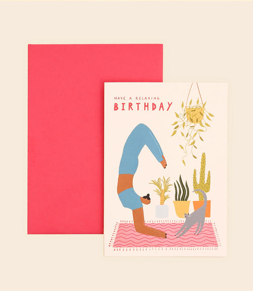 The Yoga Birthday Card by Blue Dog Posters, designed in Manchester, UK, features a person performing a yoga pose on a pink mat surrounded by pots of plants and a hanging plant. The card wishes "Have a Relaxing Birthday" in red letters and comes with a matching red envelope.