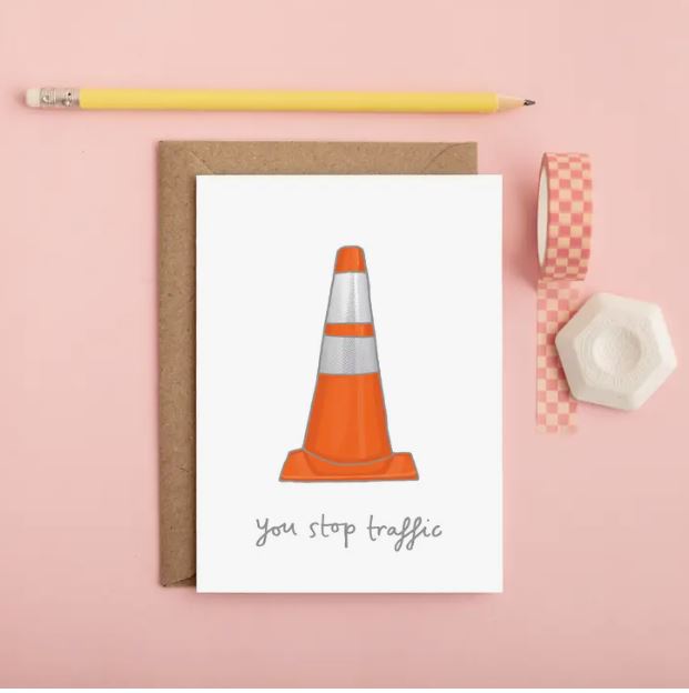 The Traffic Cone Greeting Card by Blue Dog Posters features an illustration of an orange traffic cone with white reflective stripes and handwritten words "you stop traffic" below it. The card is blank inside and comes with a brown envelope, accompanied by a yellow pencil and pink checkered tape for added charm.