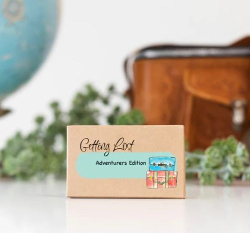 A small brown box labeled "Getting Lost Cards - Adventurers Edition" by Blue Dog Posters is depicted, featuring an illustrated image of three suitcases. In the background, there is a leather bag, some greenery, and part of a globe slightly out of focus. This travel adventure game includes misdirection cards for added excitement.