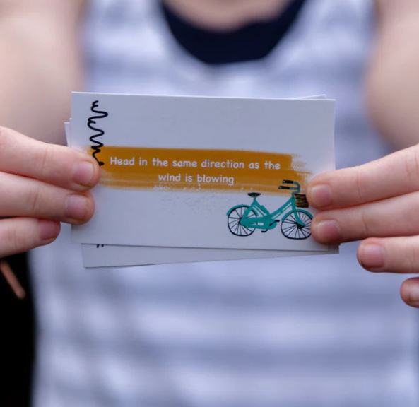 Two hands are holding a card from the Getting Lost Cards - Adventurers Edition by Blue Dog Posters, with the text "Head in the same direction as the wind is blowing" overlaying a yellow band. The card features a teal bicycle illustration on the right side, and the person holding it is wearing a striped shirt.