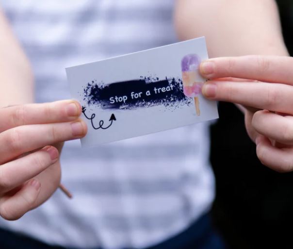 A person is holding a card from the Blue Dog Posters' "Getting Lost Cards - Adventurers Edition" with the text "Stop for a treat" and an illustration of a popsicle on it. The background is blurred, drawing attention to the card and the person's hands, dressed in a striped shirt. It feels like part of a travel adventure game—a sweet detour amidst your journey.