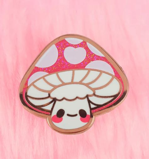 A charming accessory from Blue Dog Posters, the Amanita Mushroom Pin Pinstachio is an original illustration of a cute, smiling Amanita mushroom with blushing cheeks and big eyes. The hard enamel pin features a white cap adorned with large pink spots and glitter details, all set against a plush pink background.