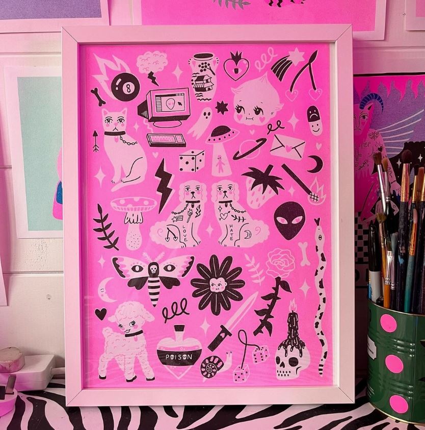 The Flash Sheet A3 Risograph by Amy Hastings, available from Blue Dog Posters, showcases a vivid pink background brimming with black and white doodles. This framed artwork features illustrations of animals, a computer, a skull, a heart, an alien face, flowers, and other whimsical designs. The frame is positioned on a desk adorned with art supplies.