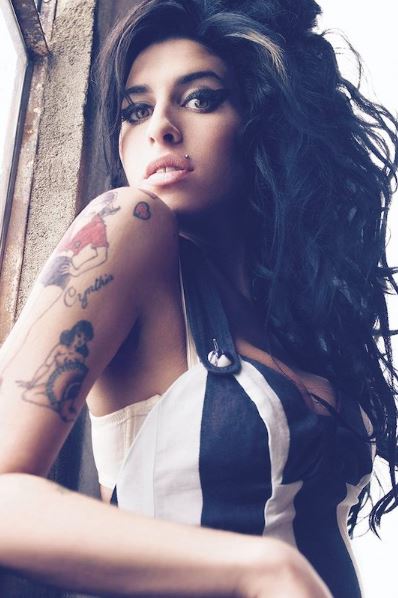 A woman with long, curly dark hair and bold eye makeup poses by a window in the MUS 024 "Amy Winehouse - Tattoos" black-and-white striped thin-strap top, showing her tattoos as light streams in beside her.