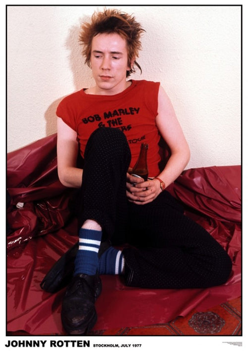 A young man with spiky hair sits on a shiny red surface against a wall. He wears a red sleeveless "Bob Marley & The Wailers" shirt, black pants, blue and white striped socks, and black shoes. Holding a bottle, he appears pensive. Text at bottom reads "Johnny Rotten, Stockholm, July 1977"—a must-have for any rock poster music collection is the **Johnny Rotten Stockholm 1977** from **MUS 282**.