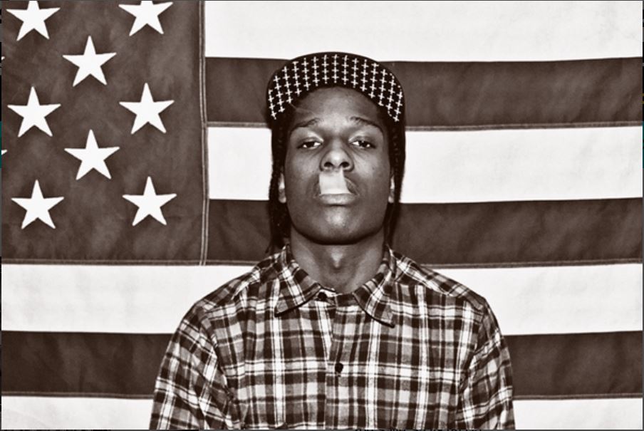 A person wearing a checkered shirt and a patterned cap stands in front of an American flag backdrop, looking directly at the camera. The black and white photo, printed on premium gloss paper, is reminiscent of a classic rap artist poster—a perfect representation of the ASAP Rocky aesthetic from the MUS 322 collection, an ideal addition to any music enthusiast's collection.