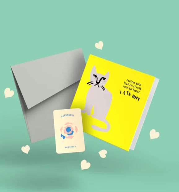 A greeting card from Blue Dog Posters titled "A Little Bird Told Me Greeting Card," featuring a cartoon cat and the text "A little bird told me it was your birthday! I ate him," sits next to a gray Colorplan Envelope. Scattered small heart shapes and an additional card with a floral design and the words "Openness Fortunes" made from plantable seed paper are also visible.