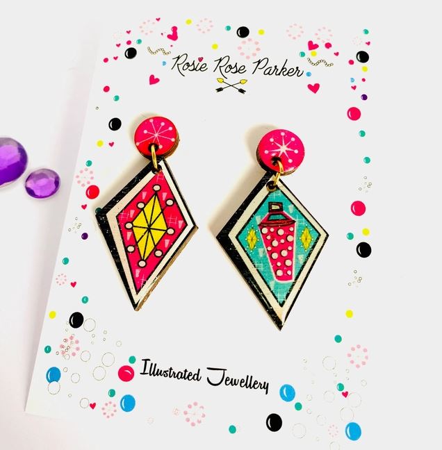 The Atomic Retro Earrings by Blue Dog Posters are displayed on a card. These colorful geometric earrings feature playful designs of a yellow diamond and a pink lantern with vibrant accents on the packaging.