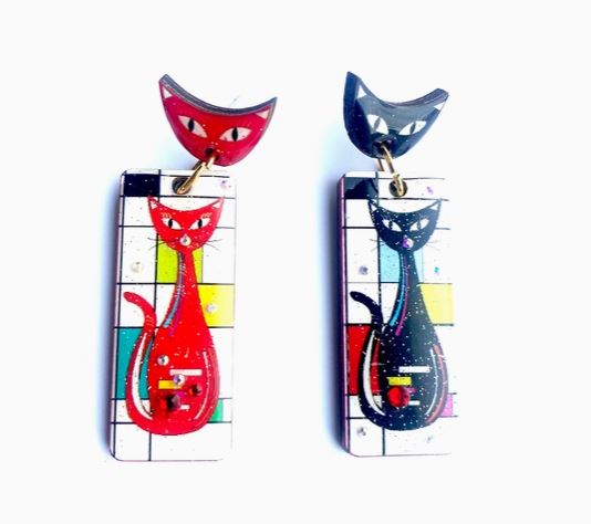 The "Atomic Cat Retro Earrings" by Blue Dog Posters feature two geometric, cat-themed earrings: each with a rectangular base and red or black cats against colorful abstract backgrounds. The tops are cat-shaped to match the cats below.