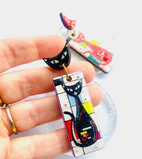 Close-up of a hand with gold rings holding a colorful rectangular Atomic Cat Retro Earring by Blue Dog Posters, featuring a black cat design inspired by Mondrian art. Another similar earring is blurred in the background.