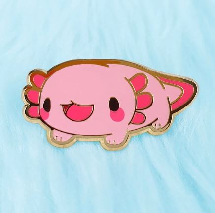 A cute Axolotl Pin Pink Pinstachiio by Blue Dog Posters with a smiling face and red cheeks lies on a light blue fuzzy surface. This adorable hard enamel pin features small legs, gill stalks, and a tail, all outlined in a golden border—perfect for fans of cute critters!