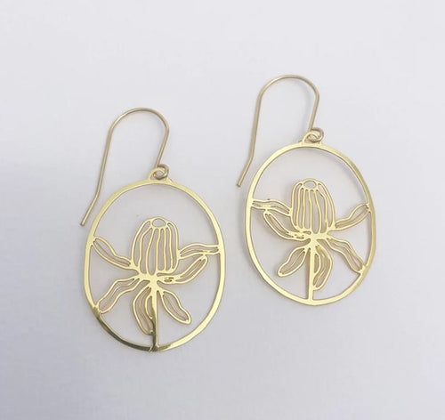 The Denz Mini Banksia Earrings Gold by Blue Dog Posters are a pair of gold earrings featuring intricate native flora designs. The floral pattern is framed within an oval shape, and each lightweight earring has a simple hook for attaching to the ear. The background is a plain, light-colored surface.